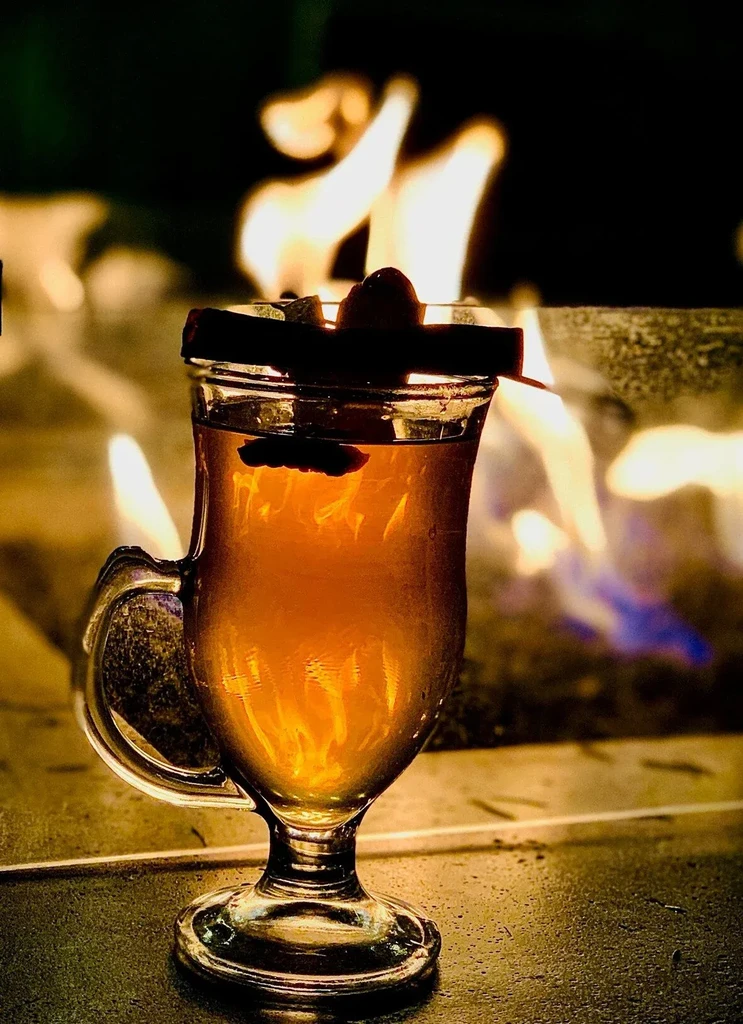 A warm drink with a cinnamon stick and cherry, with flames behind.