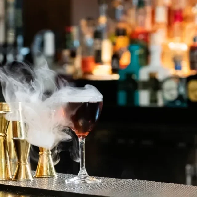 A smoking cocktail in a glass on a bar with liquor bottles.