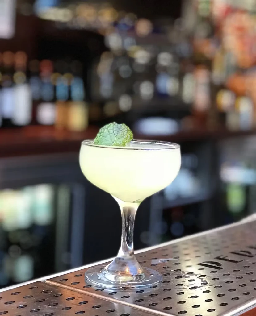 A light green cocktail with a mint leaf sits on a bar.