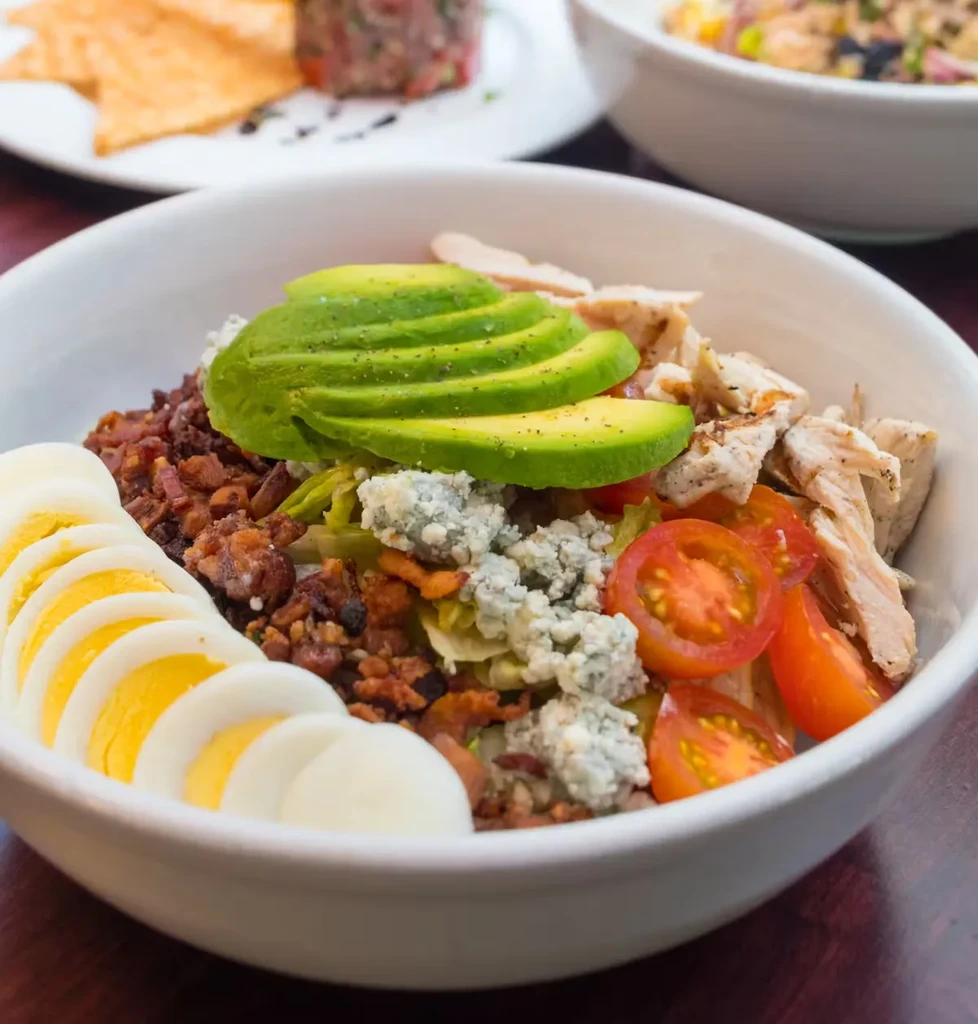 A large salad bowl with avocado, chicken, bacon, eggs, and tomatoes.