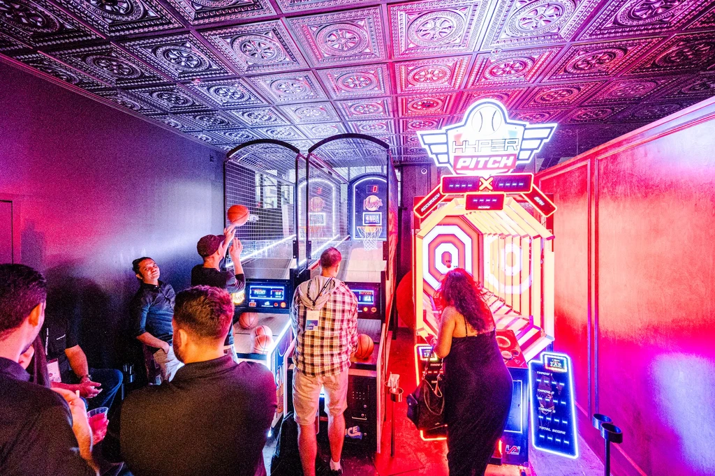 People play arcade basketball and baseball pitching games in a neon-lit room.