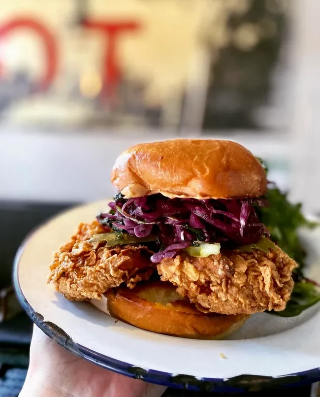 Hand holding a plate with a crispy fried chicken sandwich.