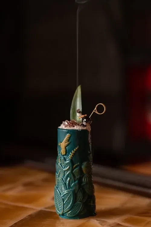 A teal tiki cocktail glass with a gold hummingbird design and smoke.