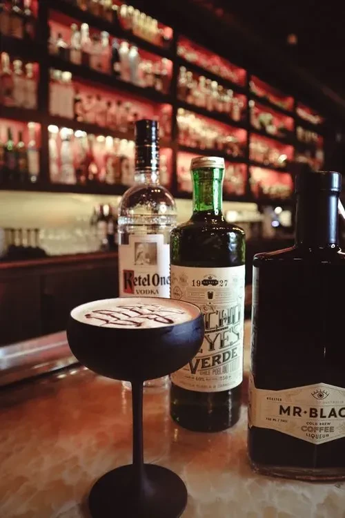 A cocktail in a dark coupe glass with three liquor bottles behind it.
