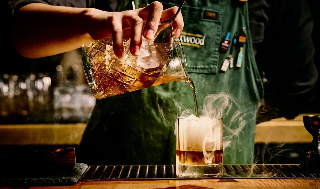 Bartender pours liquid from a patterned pitcher into a smoking glass.