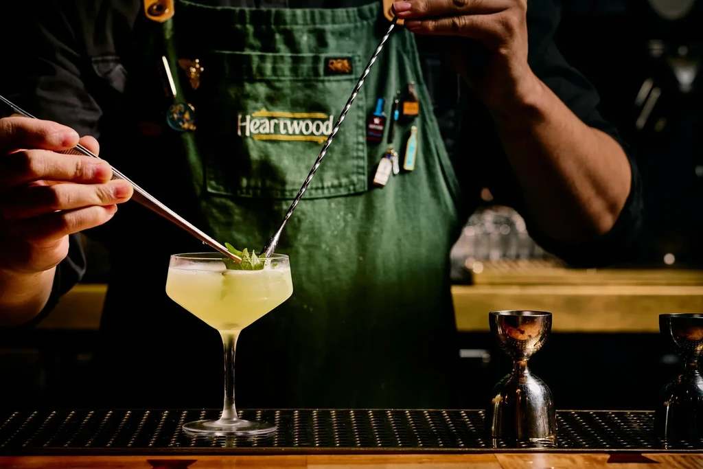 A bartender in a green apron garnishes a drink with fresh herbs.