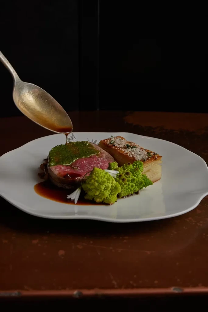 A spoon pours sauce onto a plate of meat, romanesco, and potatoes.