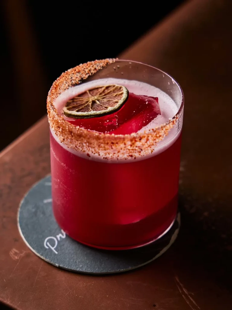 A red cocktail with a lime wheel and salted rim on a coaster.