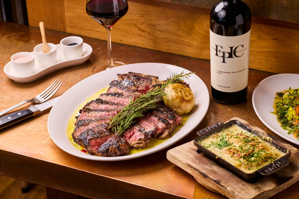 Sliced steak, roasted garlic, rosemary, and wine on a wooden table.