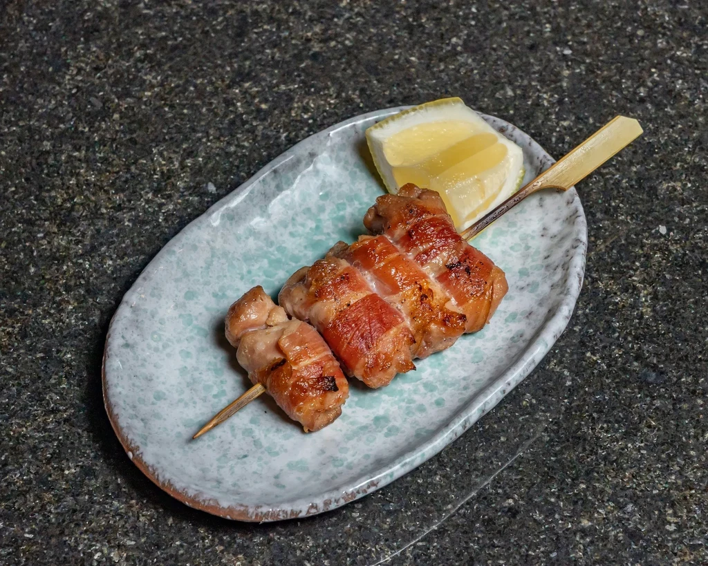 A grilled skewer with bacon-wrapped chicken and a lemon wedge on a plate.