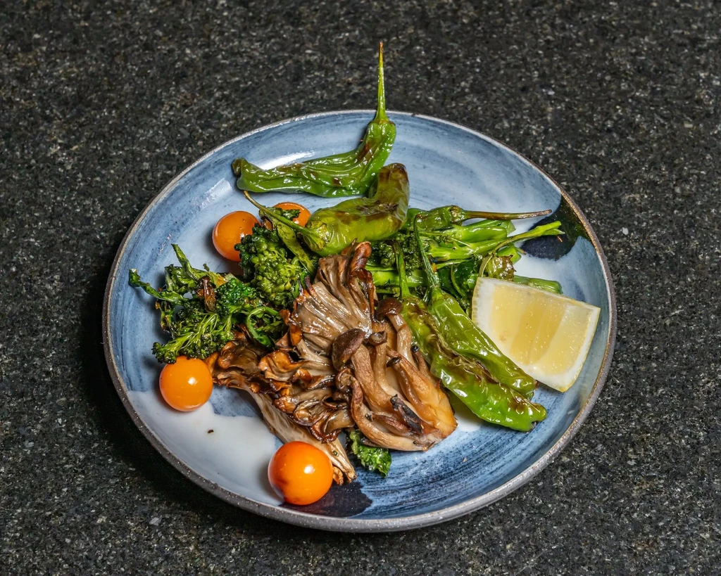 A ceramic plate with roasted vegetables, mushrooms, peppers, cherry tomatoes, and a lemon wedge.