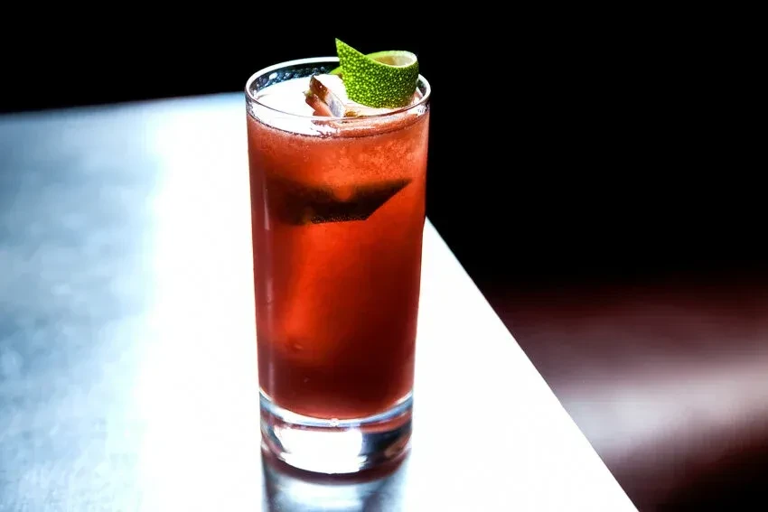 A red cocktail with lime peel garnish on a reflective table.