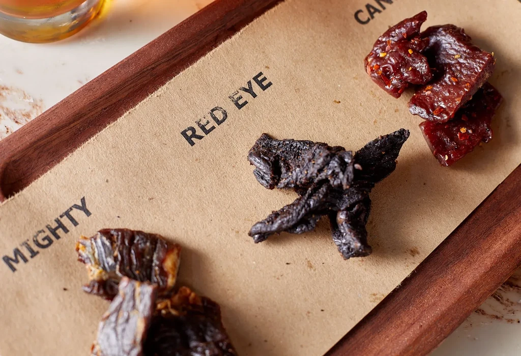 Assorted jerky pieces on a brown paper-lined wooden tray.