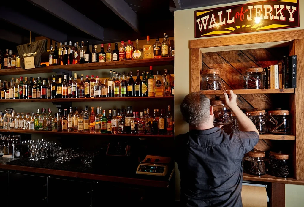 Bartender stocking liquor bottles and a "Wall of Jerky" display.