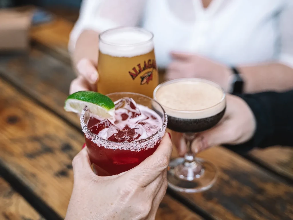 Three hands hold different drinks, cheering over a wooden table.