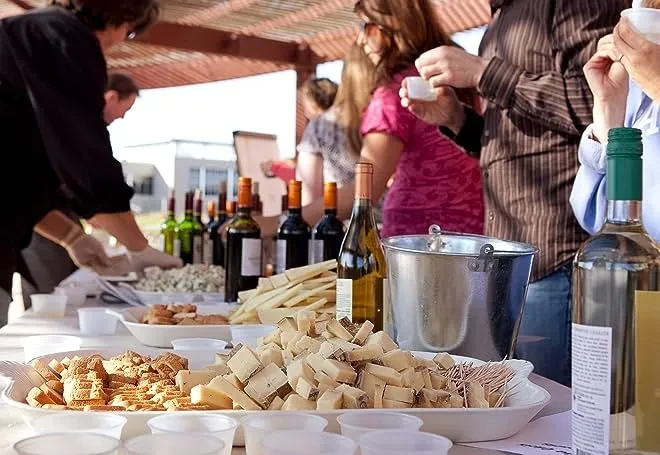 Outdoor wine tasting with people sampling snacks and various bottles of wine.