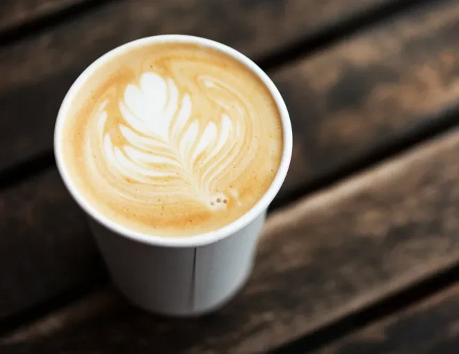 A take-out latte with leaf art in foam on a wooden table.