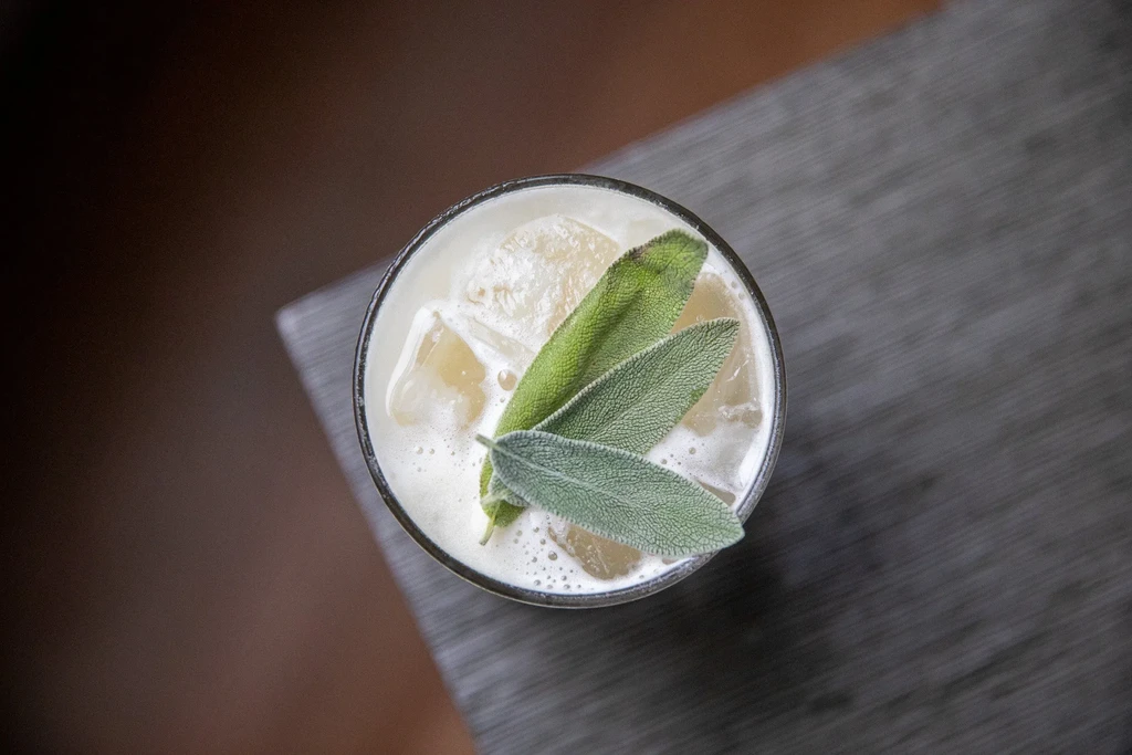 Overhead view of a glass cocktail with ice, foam, and three sage leaves.