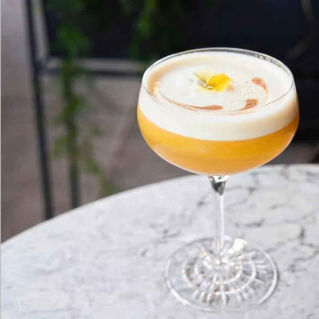 Orange cocktail with white foam, a yellow flower, and cinnamon dust.