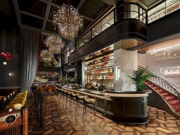 A sophisticated bar with a mezzanine, chandeliers, and red-carpeted staircase.