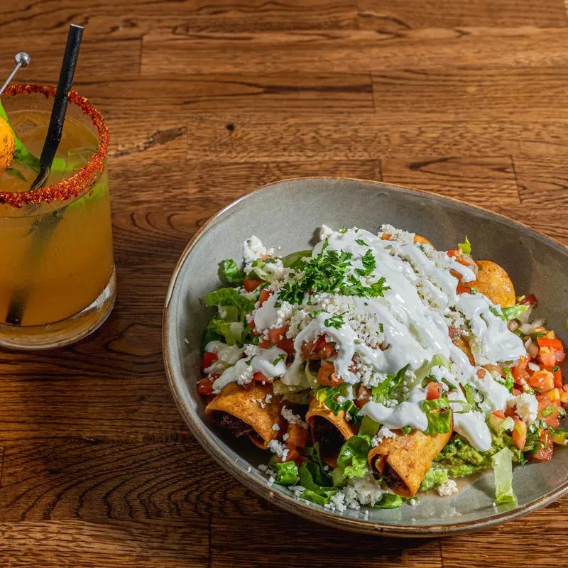 Taquitos bowl with lettuce, tomatoes, cheese, and a drink on a wooden table.