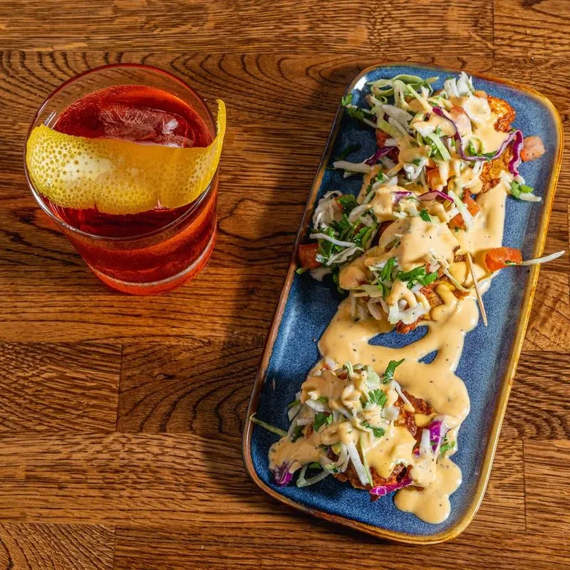 Red cocktail with lemon peel and tacos with sauce on a wooden table.