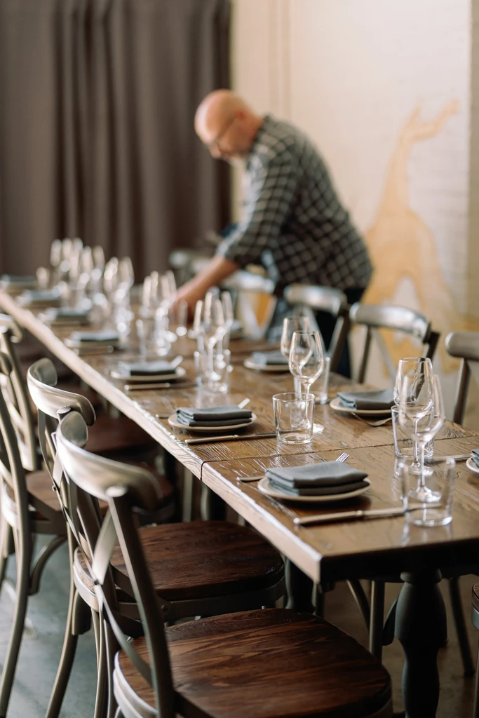 A man sets a long wooden table with dishes and glasses.