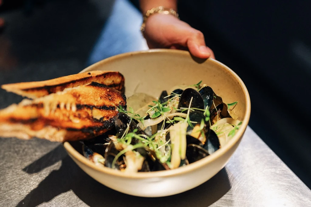 A hand holds a bowl of mussels, bread, and sprouts on a counter.