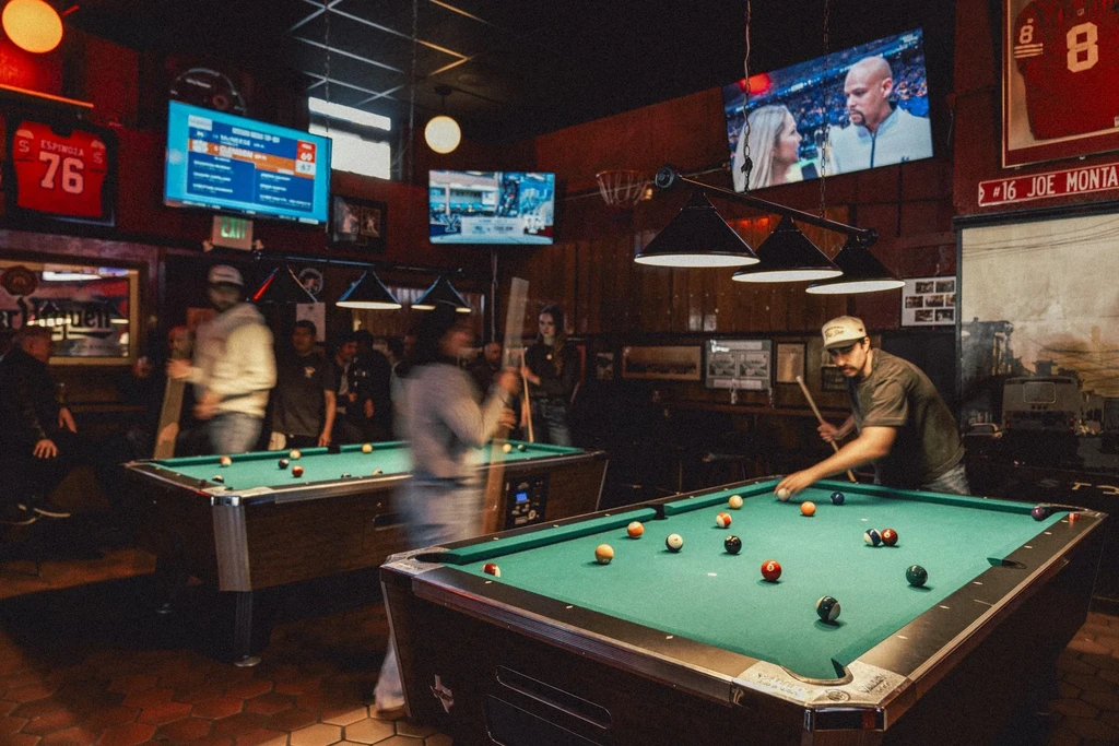 People playing billiards in a lively bar with multiple TV screens.