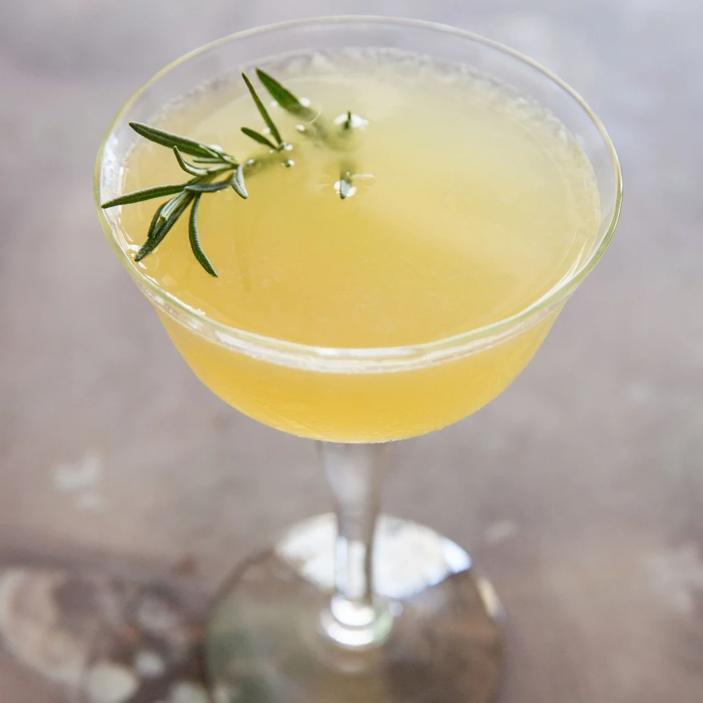A coupe glass with a yellow cocktail and rosemary garnish.