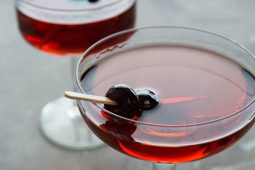 Two coupe glasses with dark red cocktails and black cherries on picks.