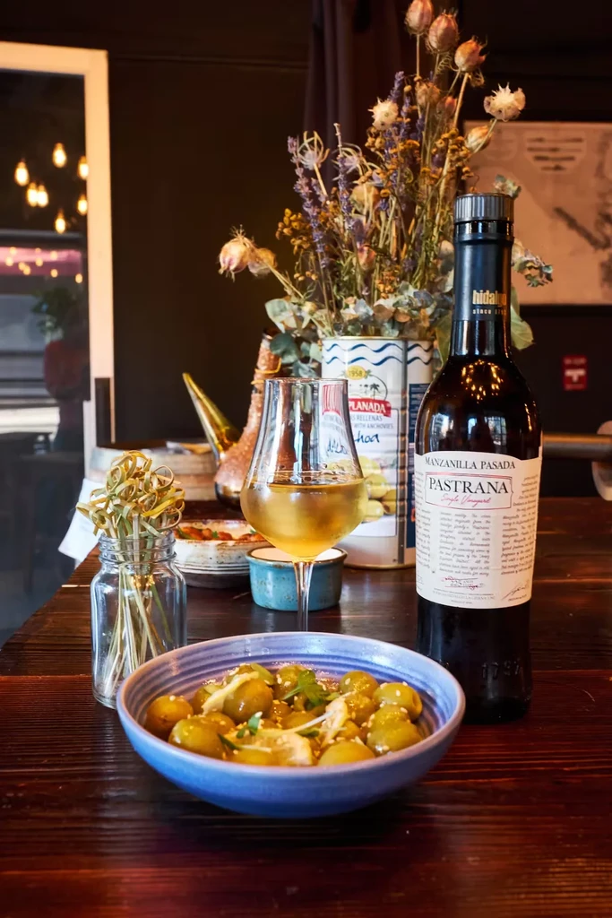 A glass of wine, olives, and a bottle of Pastrana on a table.