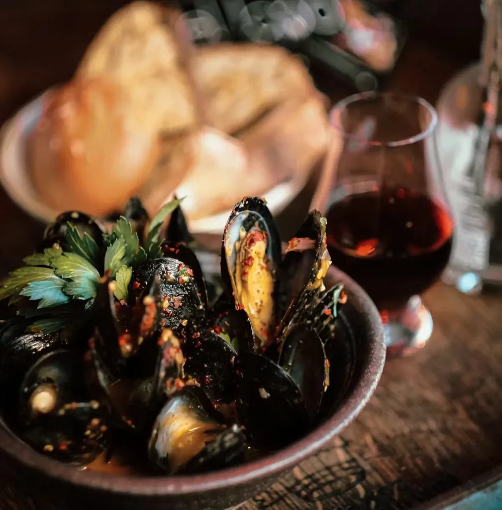 A bowl of cooked mussels with herbs, bread, and red wine.