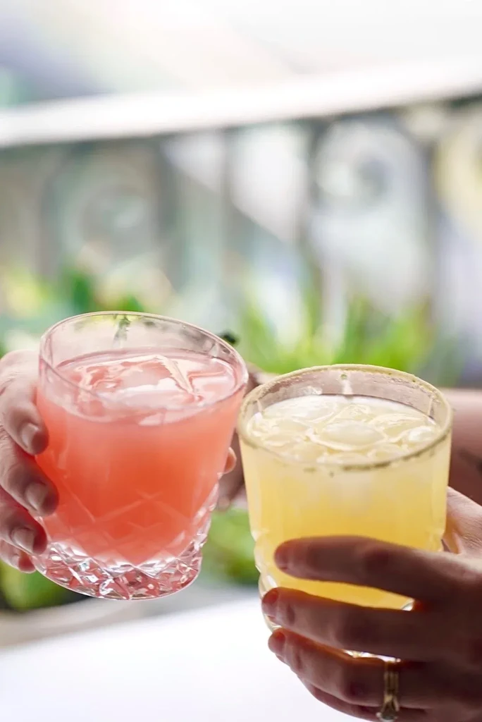Two hands holding glasses with pink and yellow iced drinks.