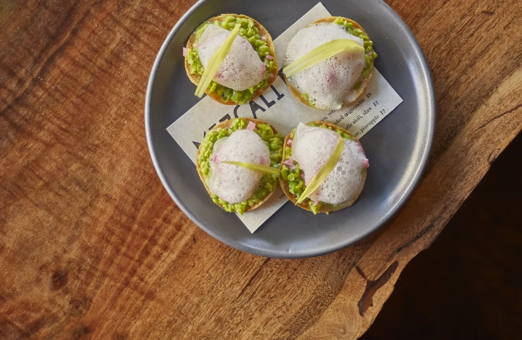 Four small tostadas with green topping, foam, and a vegetable slice.