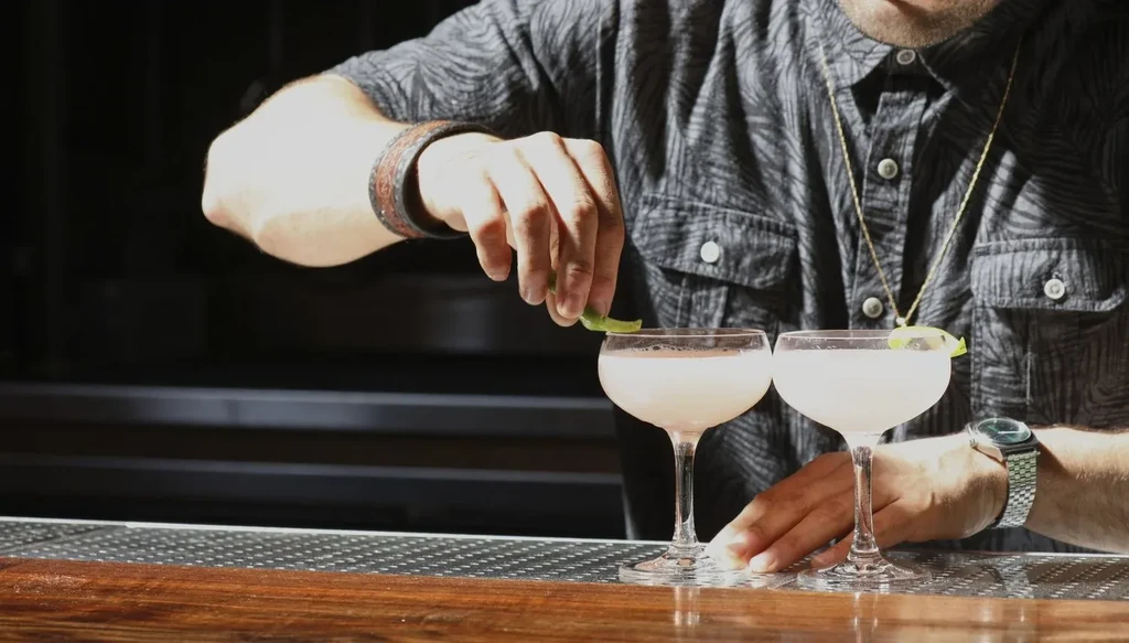 Bartender garnishes two light-pink cocktails with lime peels on a bar counter.