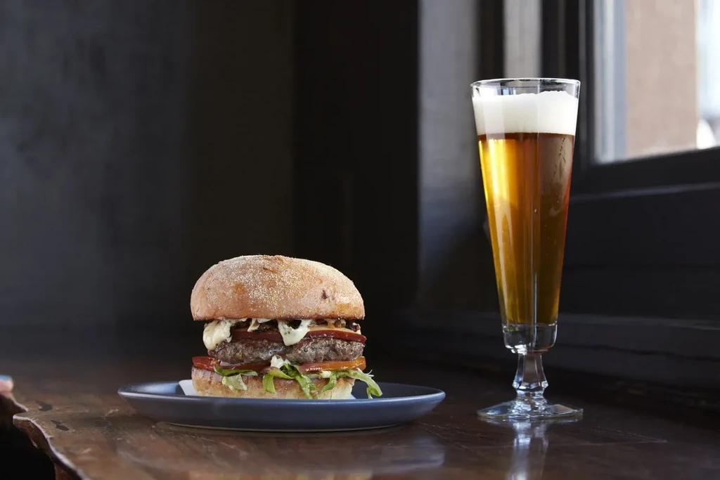 A large burger and a tall glass of beer on a wooden table.