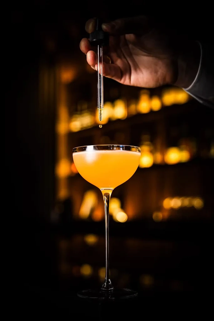 A hand drips liquid from a dropper into a yellow cocktail.