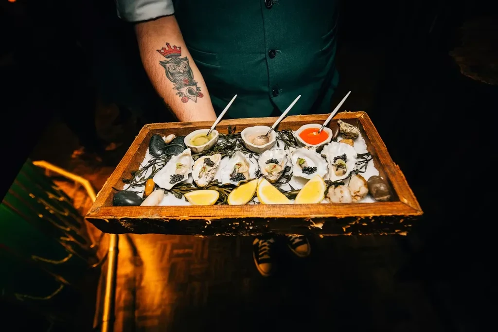 Person with owl tattoo holding tray of oysters, caviar, and lemon.