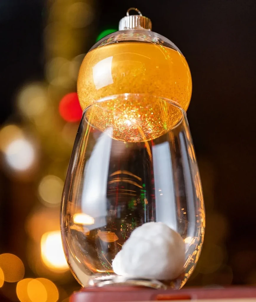 Clear wine glass holding a gold glitter ornament and cotton ball.