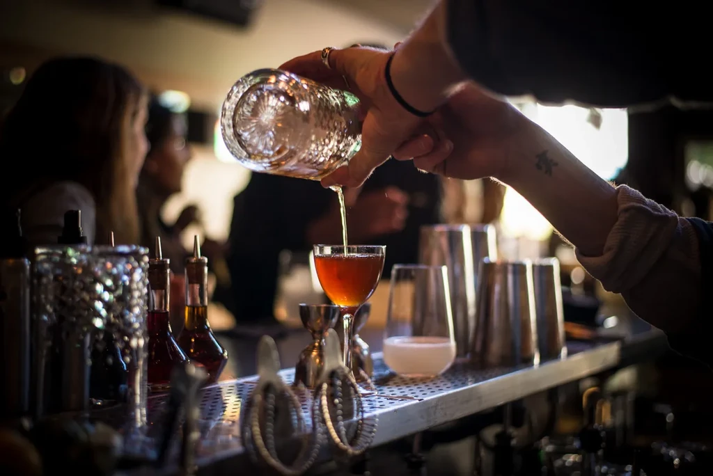 Bartender pours an amber-colored drink into a stemmed glass at a bar.