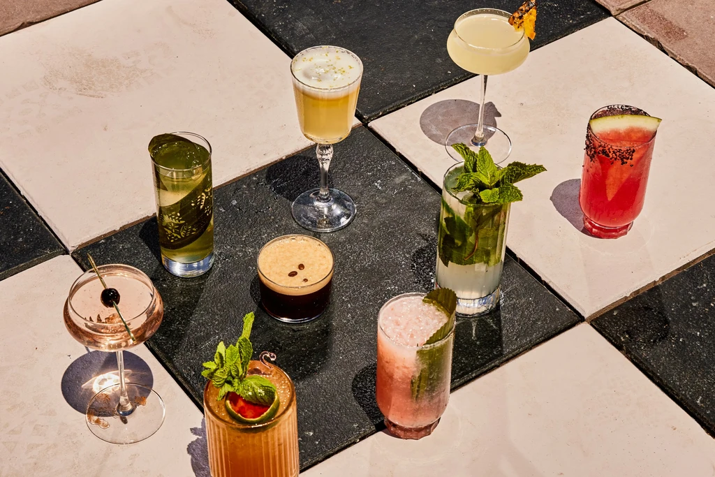 Various cocktails in different glasses are arranged on a checkered tile floor.