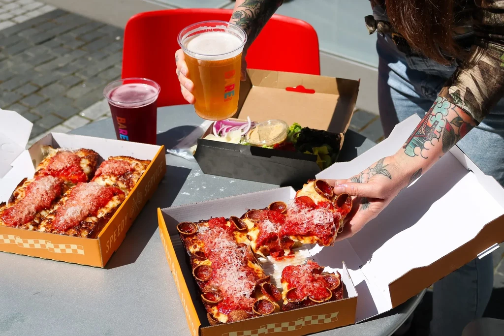 Tattooed person holding a pizza slice and beer on an outdoor table.