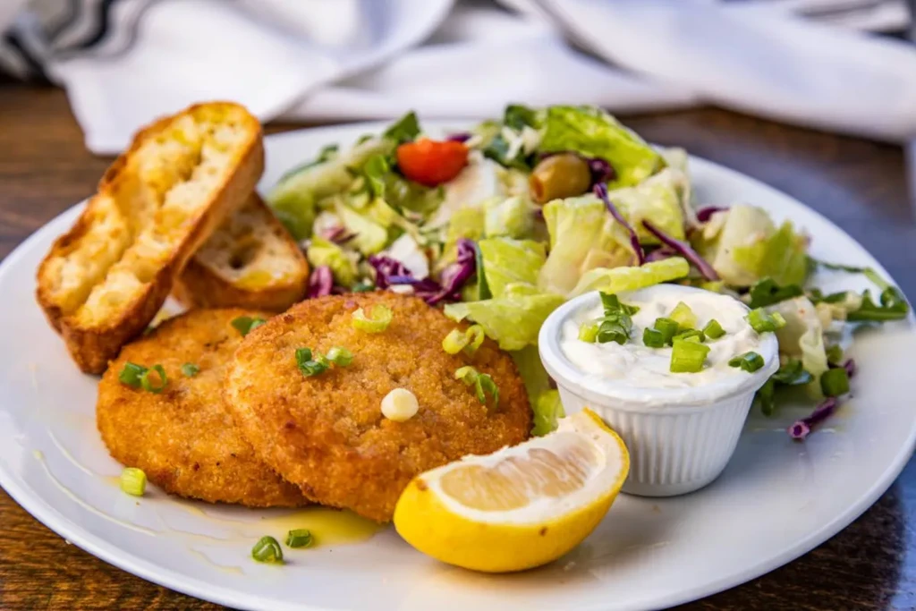 Fried patties with salad, toast, sauce, lemon slice on a white plate.