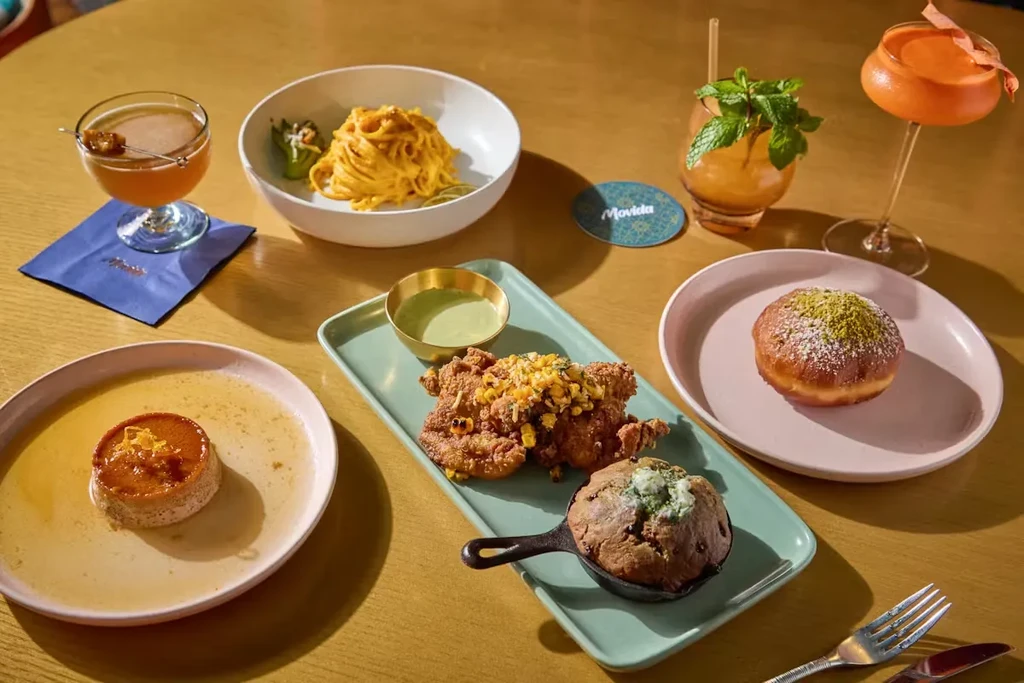 A wooden table with various dishes: pasta, fried chicken, desserts, and cocktails.