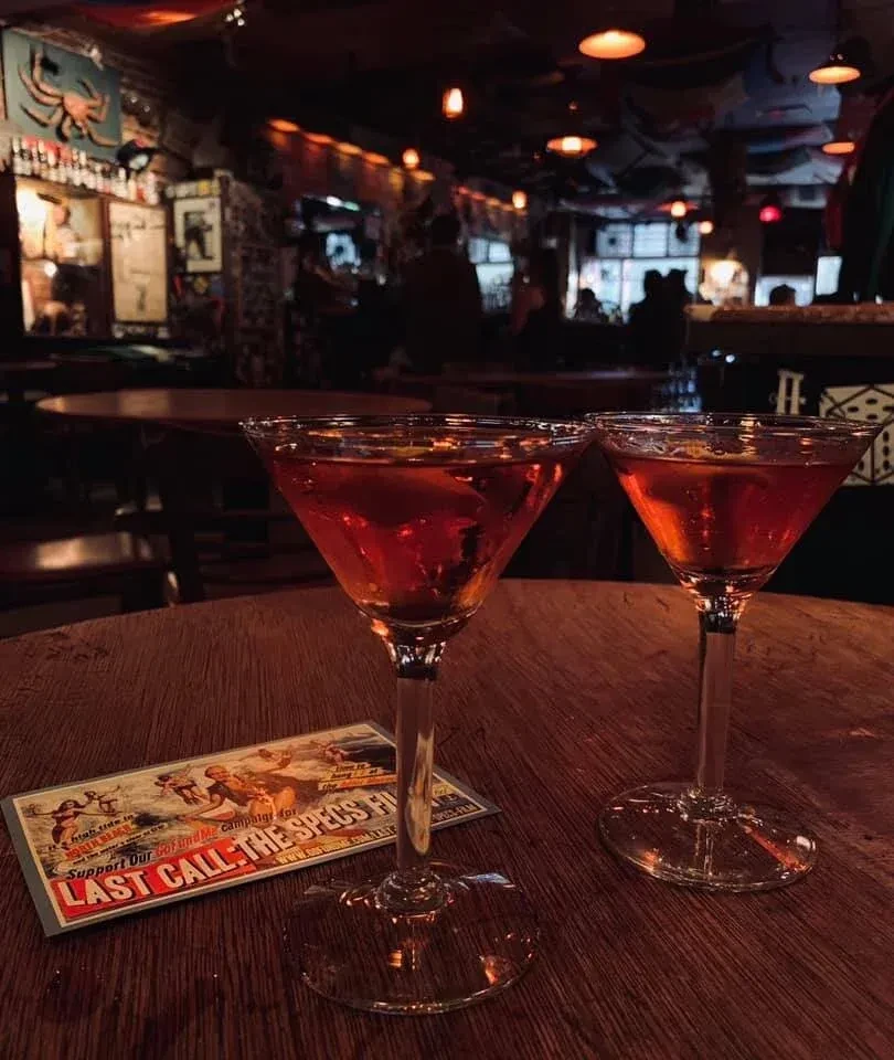 Two red cocktails in martini glasses on a wooden table in a dimly lit bar.