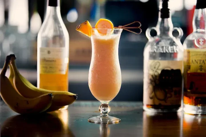 Tropical cocktail with fruit garnish, bananas, and liquor bottles on a bar.