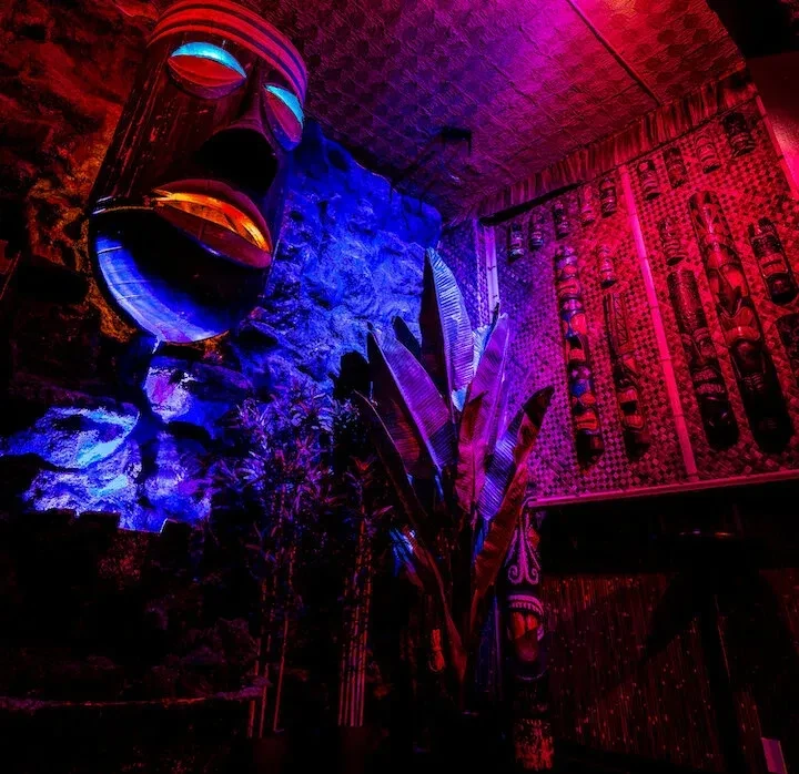 Dimly lit tiki bar with large mask, plants, and wall carvings.