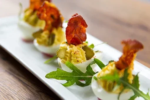 Deviled eggs topped with bacon, pickles, and green leaves.