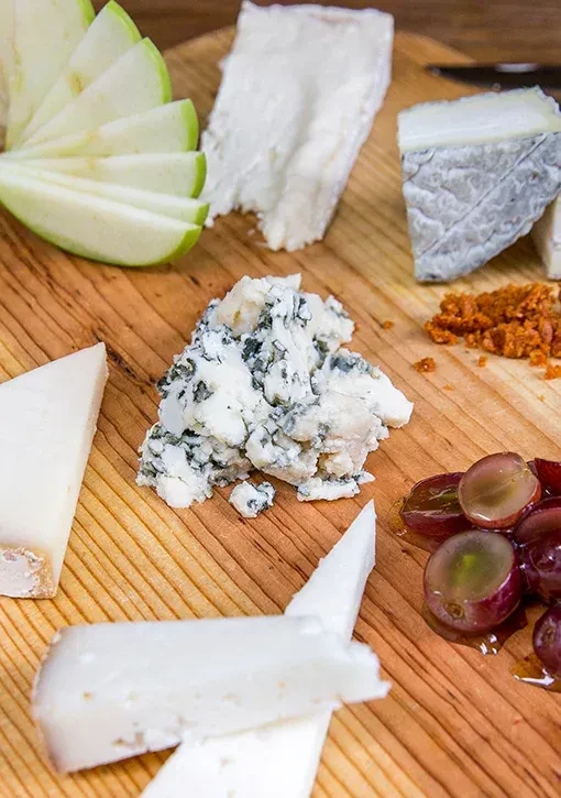 Assortment of cheeses, apple slices, grapes, and red crumbs on a wood board.